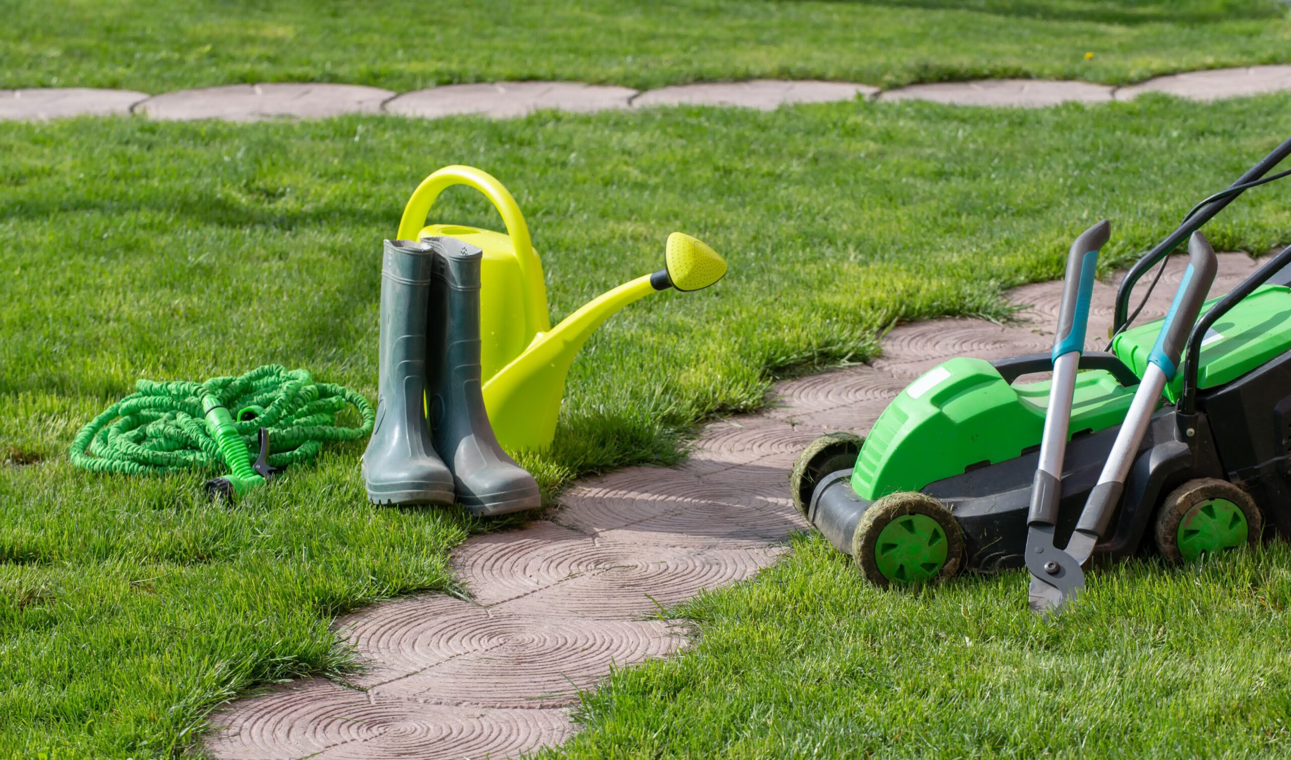 Lawn care tools including a green hose, watering can, mower, and boots on a garden path, ready for yard maintenance and watering.